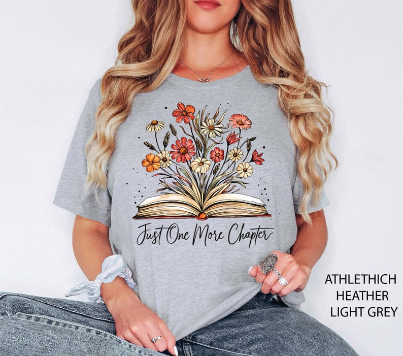 May include: A light gray t-shirt with a floral design and an open book. The text "Just One More Chapter" is printed on the shirt.