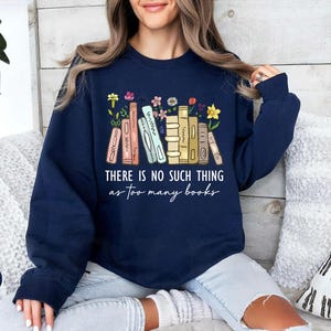 May include: Navy blue sweatshirt with the text "THERE IS NO SUCH THING as too many books" in white script. The design features a row of colorful books with flowers. The sweatshirt is a comfortable, casual style.