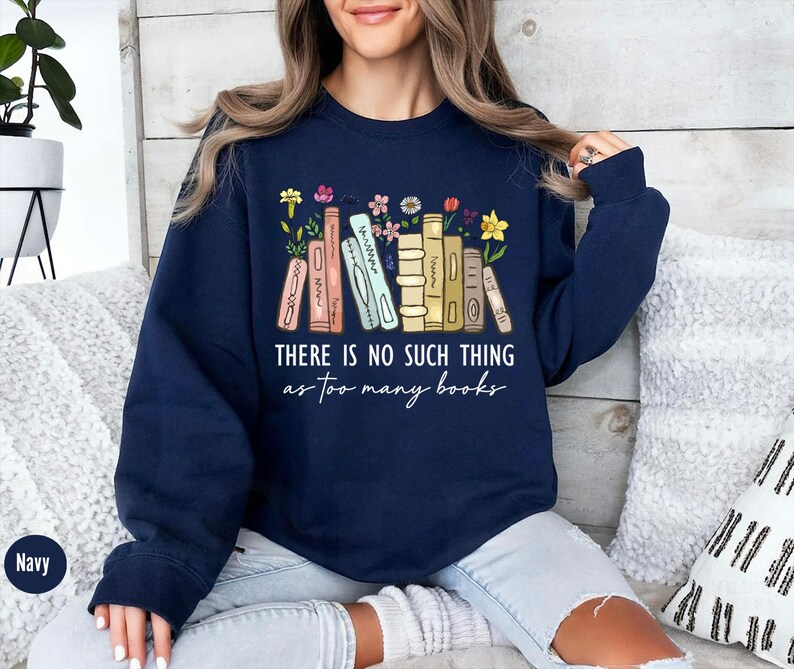 May include: Navy blue sweatshirt with the text "THERE IS NO SUCH THING as too many books" in white script. The design features a row of colorful books with flowers. The sweatshirt is a comfortable, casual style.
