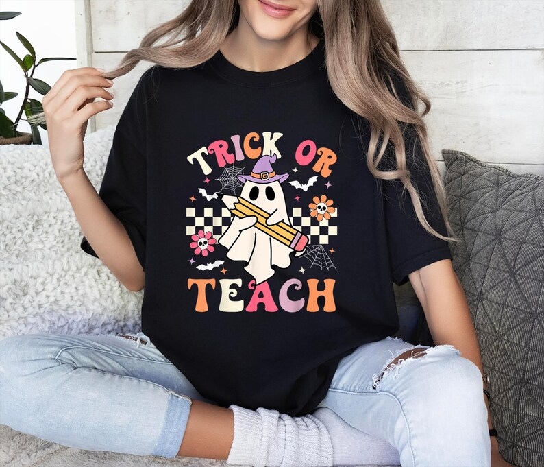 May include: A black t-shirt featuring a graphic print of a white ghost wearing a purple witch hat and holding a yellow pencil. The design includes the text "TRICK OR TEACH" in orange and pink, surrounded by spiderwebs, bats, flowers, and a checkerboard pattern.
