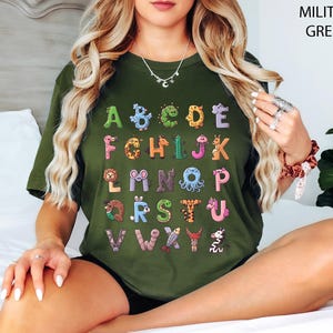 K&ouml;nnte beinhalten: Ein milit&auml;rgr&uuml;nes T-Shirt mit einem farbenfrohen Alphabet-Design. Jeder Buchstabe ist einzigartig mit Tier-Grafiken illustriert. Das Design des Shirts enth&auml;lt den Text "MILITARY GREEN" in der oberen rechten Ecke.