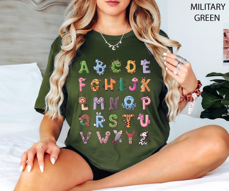 K&ouml;nnte beinhalten: Ein milit&auml;rgr&uuml;nes T-Shirt mit einem farbenfrohen Alphabet-Design. Jeder Buchstabe ist einzigartig mit Tier-Grafiken illustriert. Das Design des Shirts enth&auml;lt den Text "MILITARY GREEN" in der oberen rechten Ecke.
