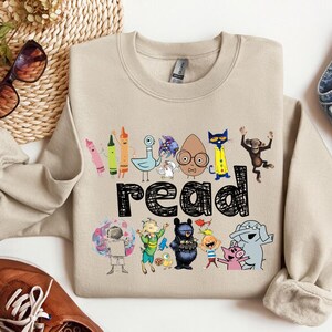 May include: Beige sweatshirt with a colorful graphic that says "read". The graphic features various cartoon characters and crayons. The sweatshirt is paired with blue jeans, brown shoes, and sunglasses.