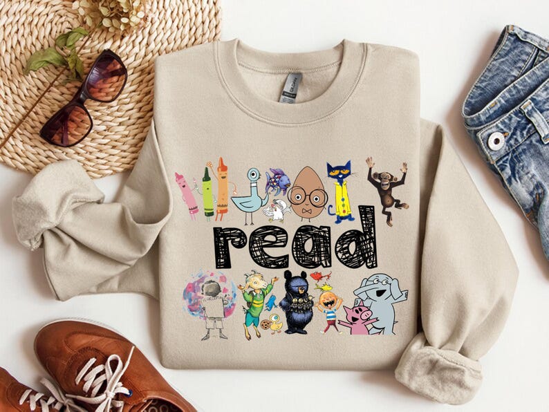 May include: Beige sweatshirt with a colorful graphic that says "read". The graphic features various cartoon characters and crayons. The sweatshirt is paired with blue jeans, brown shoes, and sunglasses.