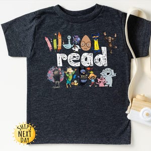 May include: Dark gray t-shirt featuring the word "read" in white, with colorful cartoon characters and crayons. The shirt has a crew neck and short sleeves. A yellow sun-shaped sticker with the text "Ships Next Day" is visible.