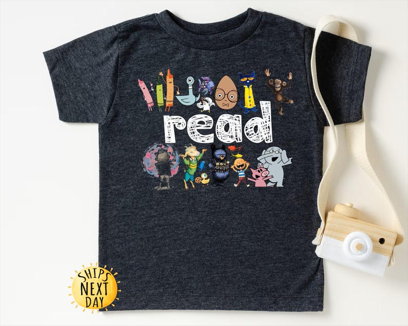 May include: Dark gray t-shirt featuring the word "read" in white, with colorful cartoon characters and crayons. The shirt has a crew neck and short sleeves. A yellow sun-shaped sticker with the text "Ships Next Day" is visible.