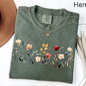 Peut inclure: Un t-shirt vert Comfort Colors avec un motif floral. Le t-shirt pr&eacute;sente une illustration de style aquarelle de fleurs et de feuilles color&eacute;es. Un collier en argent avec un petit pendentif repose sur le t-shirt. Le mot "Hemp" est imprim&eacute; en haut &agrave; droite.