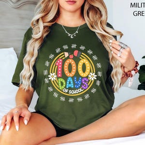 Pu&ograve; includere: Una donna indossa una maglietta verde con una grafica colorata "100 giorni di scuola". La grafica presenta un cerchio con il numero 100, le parole "Giorni di scuola" e una variet&agrave; di forme e disegni colorati.