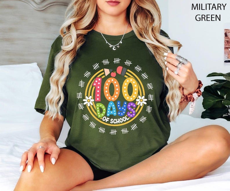 Pu&ograve; includere: Una donna indossa una maglietta verde con una grafica colorata "100 giorni di scuola". La grafica presenta un cerchio con il numero 100, le parole "Giorni di scuola" e una variet&agrave; di forme e disegni colorati.