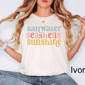 Może przedstawiać: Kremowa koszulka z napisem "saltwater seashells sunshine" w pastelowych kolorach. Koszulka ma luźny kr&oacute;j i nadaje się do noszenia na co dzień. Tekst jest w zabawnej, retro czcionce.