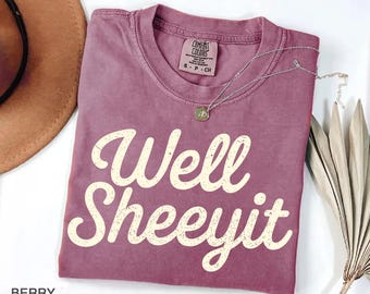 May include: A berry-colored short-sleeve t-shirt with a crew neckline. The shirt features the text Well Sheeyit printed in a distressed cream-colored font across the chest. A small tag inside the collar reads Comfort Colors size S.