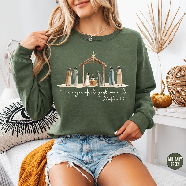 K&ouml;nnte beinhalten: Ein olivgr&uuml;nes Sweatshirt mit einer Illustration der Geburt Christi. Das Design enth&auml;lt den Text "the greatest gift of all" und "Matthew 1:21". Das Sweatshirt hat lange &Auml;rmel und eine lockere Passform.