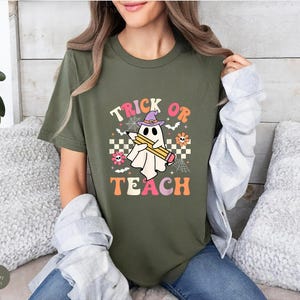 May include: A military green t-shirt featuring a graphic of a white ghost wearing a purple witch hat and holding a yellow pencil. The text reads TRICK OR TEACH, surrounded by small flowers, spiderwebs, bats, and a checkered pattern.