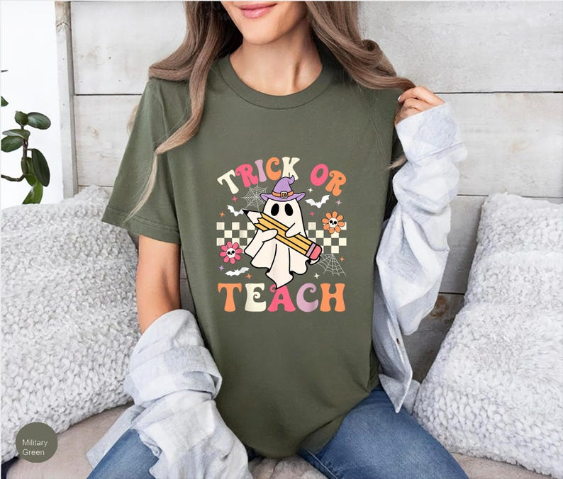 May include: A military green t-shirt featuring a graphic of a white ghost wearing a purple witch hat and holding a yellow pencil. The text reads TRICK OR TEACH, surrounded by small flowers, spiderwebs, bats, and a checkered pattern.