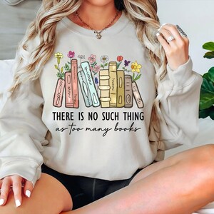 May include: Beige sweatshirt with a colorful graphic of books and flowers. The text "THERE IS NO SUCH THING as too many books" is printed below the illustration. The sweatshirt is a casual, comfortable style.