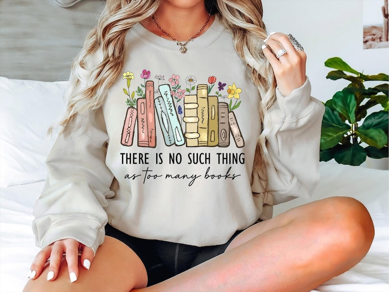 May include: Beige sweatshirt with a colorful graphic of books and flowers. The text "THERE IS NO SUCH THING as too many books" is printed below the illustration. The sweatshirt is a casual, comfortable style.