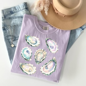 May include: A light purple t-shirt featuring watercolor oyster shell designs in shades of blue, pink, and cream. The shirt is paired with light wash denim jeans and a tan hat, creating a casual, coastal aesthetic.