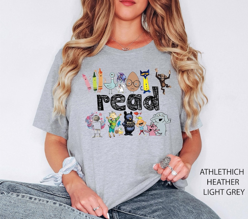 May include: Light grey t-shirt with the word "read" in black block letters. The shirt features colorful cartoon characters and crayons. The text "ATHLETHICH HEATHER LIGHT GREY" is printed on the right side.