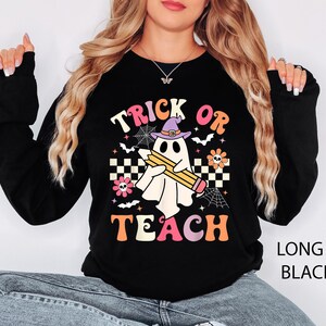 May include: A black long-sleeve sweatshirt featuring a graphic of a white ghost wearing a purple witch hat and holding a yellow pencil. The design includes the text "TRICK OR TEACH" in colorful letters, surrounded by spiderwebs, bats, and flowers.