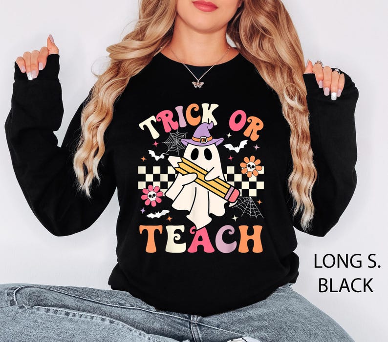 May include: A black long-sleeve sweatshirt featuring a graphic of a white ghost wearing a purple witch hat and holding a yellow pencil. The design includes the text "TRICK OR TEACH" in colorful letters, surrounded by spiderwebs, bats, and flowers.