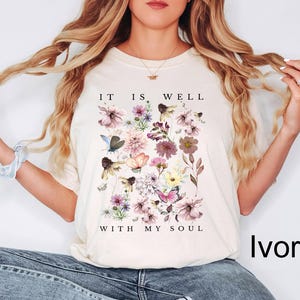 May include: An ivory short-sleeve t-shirt featuring a central graphic of various colourful wildflowers and butterflies. The text "IT IS WELL" is printed above the floral arrangement, and the text "WITH MY SOUL" is printed below the flowers.