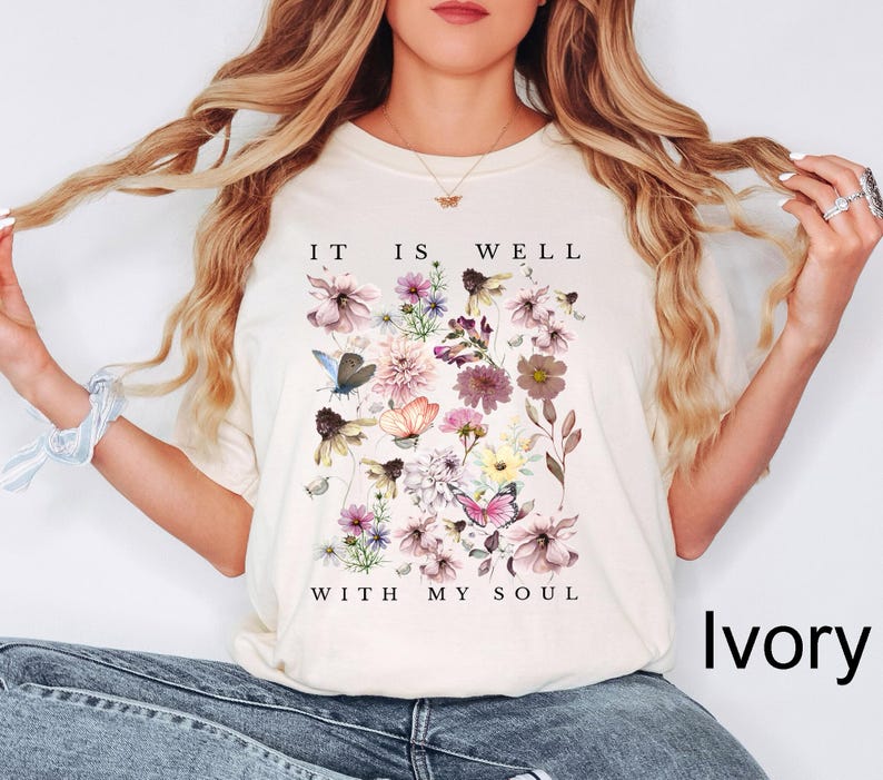 May include: An ivory short-sleeve t-shirt featuring a central graphic of various colourful wildflowers and butterflies. The text "IT IS WELL" is printed above the floral arrangement, and the text "WITH MY SOUL" is printed below the flowers.