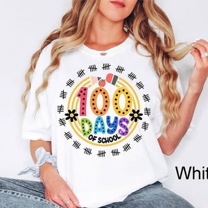 Pu&ograve; includere: Maglietta bianca con una grafica colorata che dice "100 giorni di scuola" circondata da un cerchio di segni di hash neri e un bordo arcobaleno.