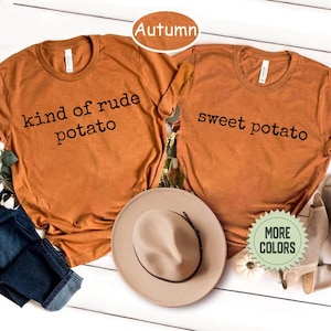 May include: Two burnt orange crew neck t-shirts featuring black text. One shirt reads "kind of rude potato" and the other reads "sweet potato." A tan wide-brim hat is positioned between the shirts, and a small green circular badge says "MORE COLORS."