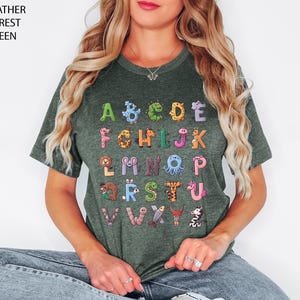 K&ouml;nnte beinhalten: Heather-Waldgr&uuml;n-T-Shirt mit farbenfrohem Alphabet-Design. Jeder Buchstabe ist mit Tierillustrationen verziert. Der Text des Shirts lautet "HEATHER FOREST GREEN" in der oberen linken Ecke.