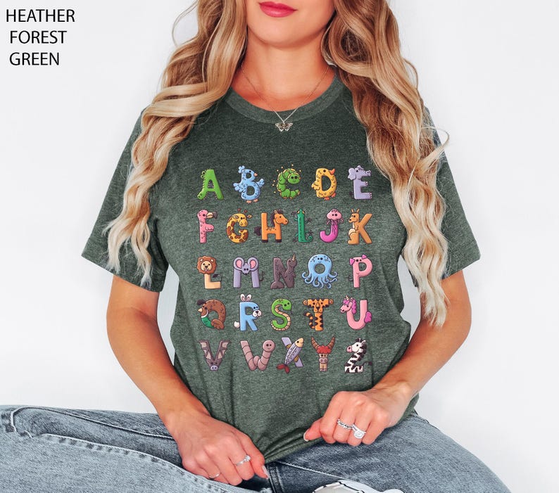 K&ouml;nnte beinhalten: Heather-Waldgr&uuml;n-T-Shirt mit farbenfrohem Alphabet-Design. Jeder Buchstabe ist mit Tierillustrationen verziert. Der Text des Shirts lautet "HEATHER FOREST GREEN" in der oberen linken Ecke.
