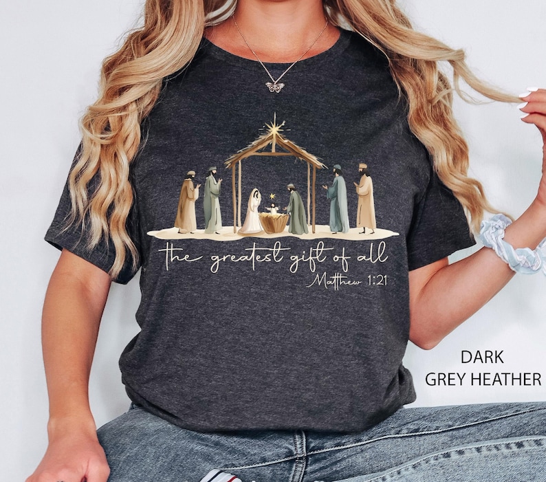 K&ouml;nnte beinhalten: Dunkelgraues Heather-T-Shirt mit einer Illustration der Weihnachtsgeschichte. Die Grafik zeigt die Heilige Familie und die Heiligen Drei K&ouml;nige. Der Text lautet "das gr&ouml;&szlig;te Geschenk von allen" und "Matth&auml;us 1:21".