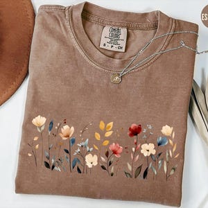 Peut inclure: T-shirt couleur expresso avec un motif floral sur le devant. Le motif pr&eacute;sente des fleurs de style aquarelle dans les tons de rouge, de jaune et de bleu. Le t-shirt a un col rond et des manches courtes. Un collier argent&eacute; avec un petit pendentif est sur le t-shirt.