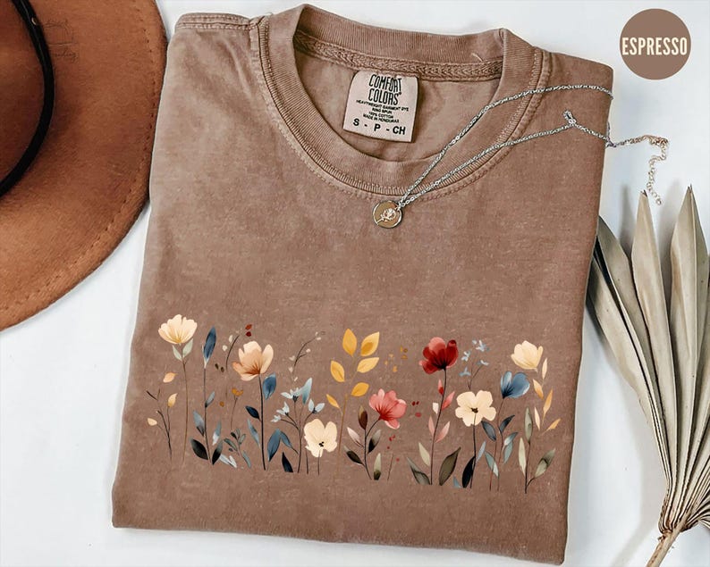 Peut inclure: T-shirt couleur expresso avec un motif floral sur le devant. Le motif pr&eacute;sente des fleurs de style aquarelle dans les tons de rouge, de jaune et de bleu. Le t-shirt a un col rond et des manches courtes. Un collier argent&eacute; avec un petit pendentif est sur le t-shirt.