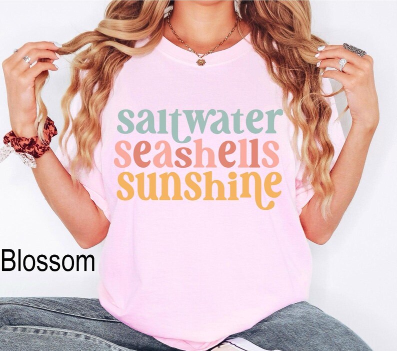 Może przedstawiać: Jasnor&oacute;żowa koszulka z napisem "saltwater seashells sunshine" w kolorze turkusowym, brzoskwiniowym, koralowym i ż&oacute;łtym. Koszulka jest w stylu casual, idealna na plażę lub lato.