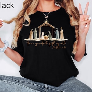 K&ouml;nnte beinhalten: Schwarzes T-Shirt mit Rundhalsausschnitt und einer Illustration der Geburt Christi. Das Design umfasst einen Stall, Figuren und den Text "the greatest gift of all" und "Matthew 1:21" in goldfarbenen Buchstaben.