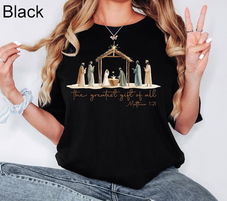 K&ouml;nnte beinhalten: Schwarzes T-Shirt mit Rundhalsausschnitt und einer Illustration der Geburt Christi. Das Design umfasst einen Stall, Figuren und den Text "the greatest gift of all" und "Matthew 1:21" in goldfarbenen Buchstaben.