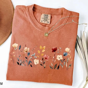 Peut inclure: Un t-shirt de couleur terre cuite avec un motif floral sur le devant. Le motif pr&eacute;sente des fleurs de style aquarelle de diff&eacute;rentes couleurs, dont le rouge, le jaune et le bleu. Un collier en or avec un pendentif repose sur le t-shirt. L'&eacute;tiquette indique "Comfort Colors".