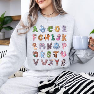 K&ouml;nnte beinhalten: Ein hellgraues Sweatshirt mit einem farbenfrohen Alphabet-Design. Jeder Buchstabe ist mit einem anderen Tier illustriert, was ein lustiges und lehrreiches Thema ergibt. Das Sweatshirt ist ein bequemer, l&auml;ssiger Stil.
