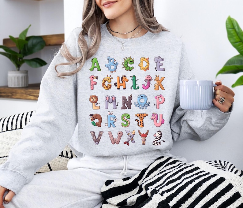 K&ouml;nnte beinhalten: Ein hellgraues Sweatshirt mit einem farbenfrohen Alphabet-Design. Jeder Buchstabe ist mit einem anderen Tier illustriert, was ein lustiges und lehrreiches Thema ergibt. Das Sweatshirt ist ein bequemer, l&auml;ssiger Stil.