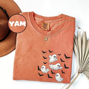 May include: Orange Comfort Colors t-shirt featuring a design of cartoon ghosts and bats. The shirt has a round neck and short sleeves. A gold necklace with a pendant is draped across the shirt. A brown hat is in the background.
