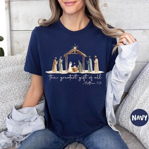 K&ouml;nnte beinhalten: Marineblaues T-Shirt mit einer Aquarellillustration der Weihnachts-Szene, mit dem Text "the greatest gift of all" und Matth&auml;us 1:21. Das Design zeigt Figuren in Gew&auml;ndern und einen Holzstall.