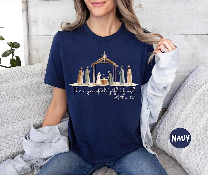 K&ouml;nnte beinhalten: Marineblaues T-Shirt mit einer Aquarellillustration der Weihnachts-Szene, mit dem Text "the greatest gift of all" und Matth&auml;us 1:21. Das Design zeigt Figuren in Gew&auml;ndern und einen Holzstall.