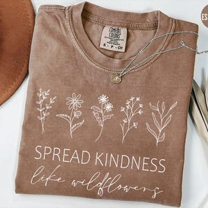 May include: An espresso-colored short-sleeve t-shirt featuring a white line-art graphic of five different wildflowers across the chest. Below the flowers, the text reads SPREAD KINDNESS in a sans-serif font and like wildflowers in script.