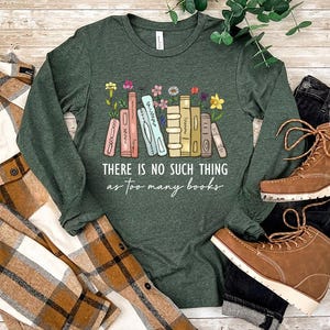 May include: Heather forest green long-sleeve shirt with the text "THERE IS NO SUCH THING as too many books" and a graphic of colorful books and flowers. The shirt is paired with a plaid shirt, black jeans, and brown boots.