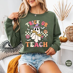 May include: A military green long-sleeve sweatshirt featuring a graphic of a white ghost wearing a purple witch hat and holding a yellow pencil. The text reads TRICK OR TEACH, surrounded by small bats, spiderwebs, flowers, and a checkerboard pattern.