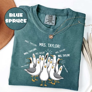 May include: A blue spruce colored t-shirt featuring a graphic of multiple seagulls with open beaks and the repeated text "MRS. TAYLOR!" surrounding them. The shirt has a crew neckline and a Comfort Colors brand label printed on the inside collar.