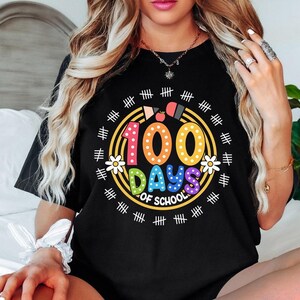 Pu&ograve; includere: Maglietta nera con una grafica colorata che dice "100 giorni di scuola" circondata da segni di hash. La grafica presenta un numero 100 arcobaleno con pois bianchi, una mela rossa e fiori gialli.