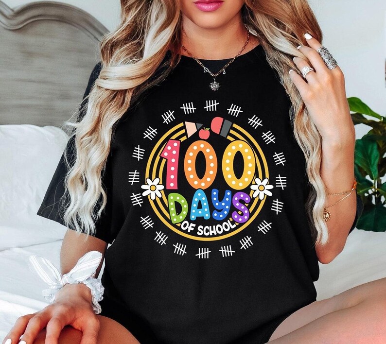 Pu&ograve; includere: Maglietta nera con una grafica colorata che dice "100 giorni di scuola" circondata da segni di hash. La grafica presenta un numero 100 arcobaleno con pois bianchi, una mela rossa e fiori gialli.