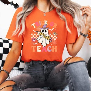 May include: An orange short-sleeve t-shirt featuring a graphic of a white ghost wearing a purple witch hat and holding a yellow pencil. The text "TRICK OR TEACH" is printed in colorful letters, surrounded by small flowers, spiderwebs, and bats.