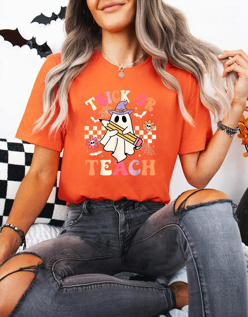 May include: An orange short-sleeve t-shirt featuring a graphic of a white ghost wearing a purple witch hat and holding a yellow pencil. The text "TRICK OR TEACH" is printed in colorful letters, surrounded by small flowers, spiderwebs, and bats.